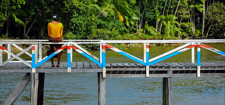Fotografia mostra uma ponte de madeira, com as laterais pintadas nas cores branca, azul e vermelha. Em baixo da ponte, passa um rio. Sentada na proteção lateral, de costas para nós, uma mulher observa a vegetação no fundo da imagem. 